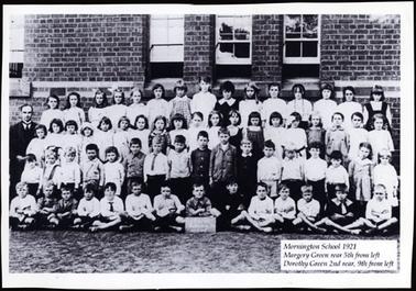 This image shows a group on 64 children arranged in four tiers. The male teacher is standing on the left of the photo, he is suited with a high collar shirt and tie. In the back ground is the school which is built of brick, and three sash windows are visible. The front row of children are sitting cross legged on the ground. 