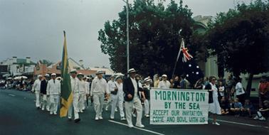 Australia Day parade in Main Street showing a group of men and women dressed in white uniforms from Mornington Bowling Club marching past the Mechanics Institute. A man and woman in front of the group are bearing a white banner with words in green “Mornington by the Sea  Accept Our Invitation  Come and Bowl With Us”. In the background a group of spectators can be seen lining the roadside.