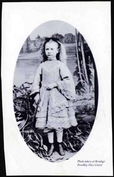 This is a studio photo of a pretty young girl. She is wearing Victorian dress. It has frills around the hem and sleeves. Her hair is long and blonde, and  she has a black bow on the top of her head. She has black ankle boots and white tights. It is oval shaped. The back ground is obviously painted landscape.