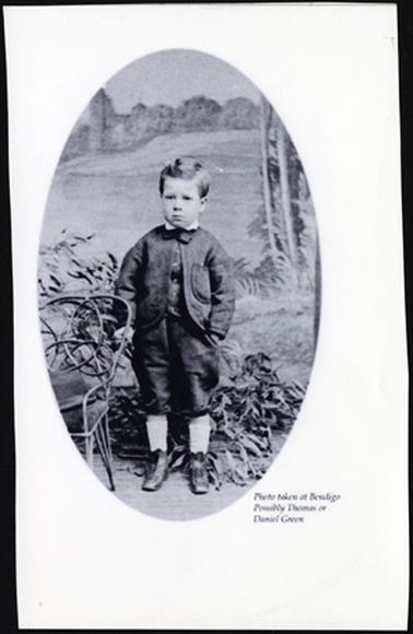 This image shows a smartly dressed young boy. He appears to be 5 or 6 years old. He is wearing a suit with a waistcoat and  short trousers, and a bow tie. His hair is smoothly combed with a side part. He has boots, and white tights. It is taken in a studio and there is a painted landscape background. It is an oval shaped photo. There is cane chair on the left of the photo.