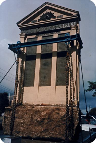 A large grey/beige concrete and bronze structure over 3 metres tall and and nearly 2 metres wide,standing on what appears to be a rock (granite?) base. The title ‘The Mornington Honour Roll’ in bronze above the three columns of names is topped by a concrete gable emblazoned with ‘1914, the rising sun badge 1918’, all in bronze. There are 121 names on the memorial. The structure has been removed from Elliott square on the south side of the Mechanics Institute and hoisted on a truck tray, secured with chains and metal girders, for transport to Memorial Park, Barkly Street, Mornington.