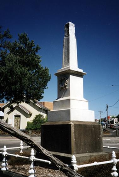 This photo shows the Football Memorial from the Beach side, with the Court House in the background. It is a stone obelisk, and has a small square garden around it, with a wrought iron fence painted white.
