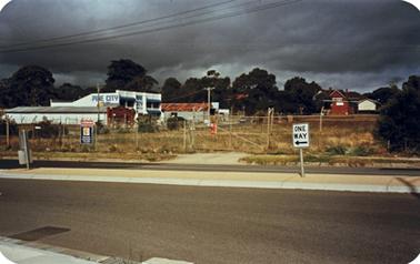A collection of timber buildings with an entrance off a main road. The driveway has a gravel surface and the facade of the main building is signed FREAN & RIDE  with Cabot’s WOOD STAINS below that. A vertical sign on the left is marked HARDWARE and a further sign on a tall pole says Frean and Ride Hardware and Timber. A sign on a wire fence reads ‘Drive in Timber”A sandwich board announces ‘OPEN 7 Days’. A yellow utility truck stands in the yard. 
