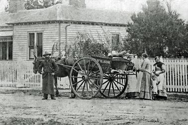 This shows a horse and cart in front of a weather board house with a picket fence. There is a bearded gentleman next to the horse with his hands on his hips. He is wearing a hat and a striped apron. There are three ladies in Victorian dress and a small child at the rear of the cart. On the side of the cart there is the name “Wilson Butcher”.