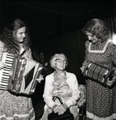 Three female figures, one seated in the middle, the other two playing accordions.