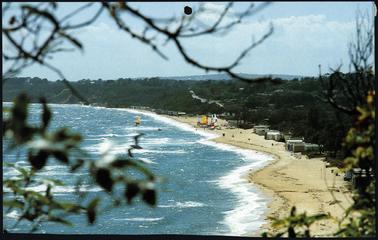This is a sweeping view of Mount Martha beach taken from above, and looking down. The sea is on the left of the photo, and the beach on the right. There are many bathing boxes visible, and in the centre of the photo are several yachts on the beach. It is a sunny day, and the sea is very blue. Only one yacht can be seen on the water.