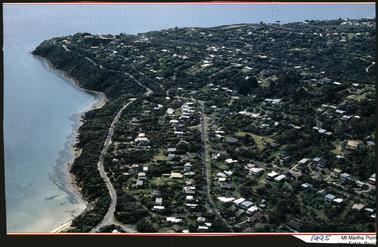 This is a view of Mt Martha from above. The sea is on the left of the photo, and the land on the left. It is an aerial view. It is quite built up, with many roofs visible. There is a lot of greenery visible, and many trees.