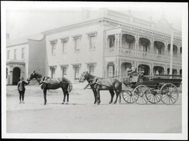 This photo shows the Royal Hotel with horses and cart in front. The Royal Hotel in the back ground has beautiful iron work to both verandahs, up and down. There are 3 black horses pulling the cart which is stationery. There is a man stood holding the reins of the leading horse, and another man sitting in the driving seat. The cart appears to be a passenger carrying vehicle. 