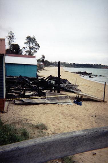 This image shows the beach, and in the centre of the image are the burned remains of a Bathing Box. Behind that is another Bathing Box painted Blue with a white roof and it is unharmed. The sea is on the right of the photo. In the foreground there is the top rung of a fence. The burned area has been cordoned off with tape.