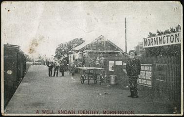 Image shows steam train to left of photo, four people on platform, one a well known identity, 
advertising hording on station fence