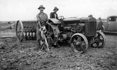 A seed driller attached to a tractor with an unidentified man in the tractor driver’s seat and another man and a dog standing beside. In the background is a man beside an utility truck.