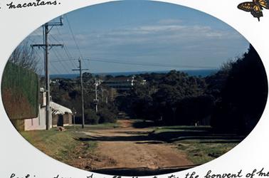 Oval shaped image of a dirt road with trees and vegetation along the verge and a white coloured house on the left and a series of telegraph poles and lines. In the distance is a view of the bay and St Macartan’s school and the Convent of Mercy