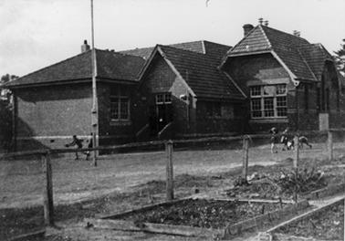 A brick school building in turn-of-the-century-style, photographed from the front. There are three rooms set behind one another and the roof of another room is just visible behind. The tiled roofs are high pitched and there are two chimneys visible. A flagpole stands in the featureless school grounds. Five or six school children are playing in two groups. A post and rail fence, with two strands of wire between the posts, defines the playground in front of the school.