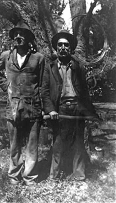 Two men in rough work clothes standing in front of a tree. The man on the right is holding an axe and a roll of wire, possibly fencing wire, hangs from a branch of the tree.