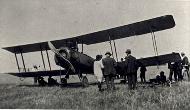 A biplane with a single engine is parked on a paddock. There are about a dozen men attired in suit and hat plus one woman dressed in a light coat, trousers and a hat. Most are standing and a few seated on the ground.