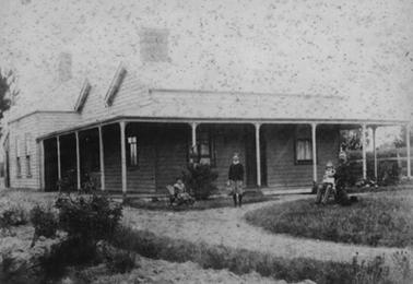 Single storied timber house in Federation style with metal double pitched roof and verandah. There is a sashed window on each side of the front door and more windows on the left. The verandah runs on the left side to a room at the back of the house. A lean-to at the back is just visible. Alfred Downward is seated on the right with daughter Ivy on his knee, Herbert is standing in the centre and Elsie is seated on the left.
