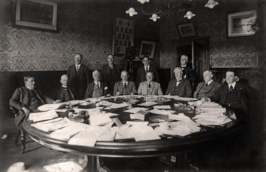 Twelve men, eight seated and four standing behind them, are around a large table covered in papers. Alfred Downward is the fourth on the right of the standing men. The table stands in an imposing room with dark wood panelling on the lower half of the walls topped by decorative wall paper.