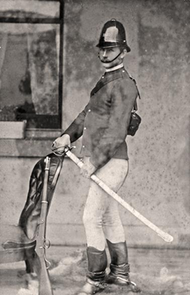 Photograph of an upright, handsome young man in police trooper’s dress of jacket, close fitting trousers, boots and a policeman’s helmet. He is holding a sword at an angle at mid-thigh and  his right hand is resting on the back of a chair on which leans a rifle. The photo appears to 