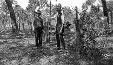 Two men in work attire, including hats and waistcoats, stand in front of a wooden gate between scrubby paddocks