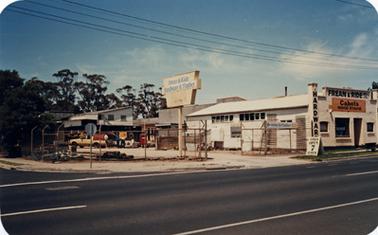 A scene with a view across Barkly Street Mornington  to cleared, vacant land covered in dry grass. to the left is a collection of commercial buildings with a prominent white building labelled ‘PINE CITY’. There are two non-descript buildings to the rear of the cleared land and a row of mature trees in the background.