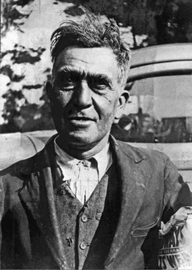 Photograph (head and shoulders) of a fine looking, tough working man dressed in full suit of workclothes. Part of motor vehicle visible behind him.