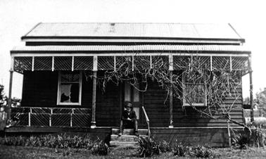 Old double fronted dark weather board house with corrugated iron roof and bull-nosed verandah. A man is seated on the front verandah with his feet on one of three steps to the front yard.