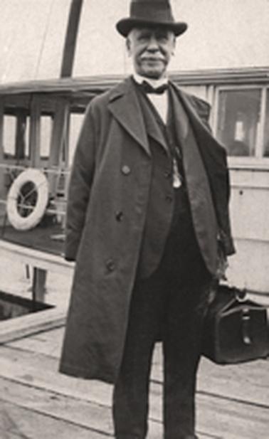 Well-dressed elderly man in hat and overcoat, carrying a gladstone bag. He stands on a wooden jetty in front of a ferry.