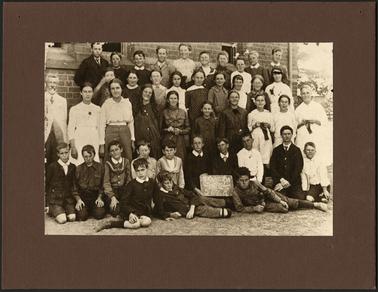 This shows a class of 41 pupils, both boys and girls. They are ranged in four tiers, with 3 children lying in the front on the grass. There are 2 teachers shown, a man standing on the left and a woman standing on the right. A child in the front row is holding a sign, it states “Mornington School Grades 5,6,7 & 8. The brick building in the background is the school.