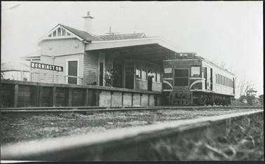 The image is of the Mornington Railway Station with four passengers waiting on the platform, to the left of the image is the ‘MORNINGTON’ sign, the engine shown in the picture was built in 1938