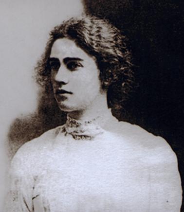 Head and shoulders portrait of a young woman in a simple high necked dress. She has a head of thick, curly hair.