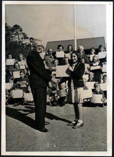 This image shows a group of children ranged in four tiers, each is holding a certificate in front of them. There are two men presumably teachers in the group. In the foreground is a gentleman presenting a certificate to a young girl. He has white hair, and a dark suit, and she is wearing a school dress and blazer. They are both smiling. There is a flag pole behind the children.