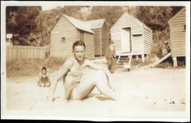 This photo shows a man in bathers, sitting on a beach in the centre of the picture. On the left of the photo is a young boy sitting on the sand. A man in a suit can be seen behind them both. There are a number of bathing boxes on the beach in the back ground.