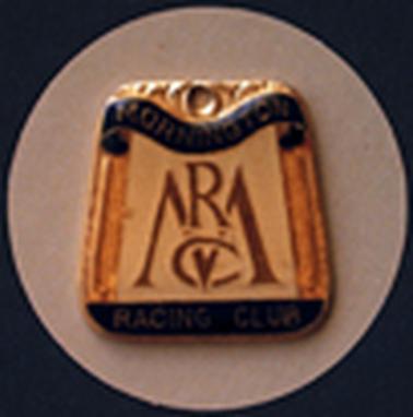 A rectangular metal badge, wider at the bottom, has a gold pattern at the top, ‘Mornington’ on a curved blue ribbon below it and ‘Racing Club’ along the black strip at the bottom. In the centre is ’MRC’ in golden letters on a beige background, with a golden whip on either side of the monogram. The badge is photographed on a fawn  coloured circular background.