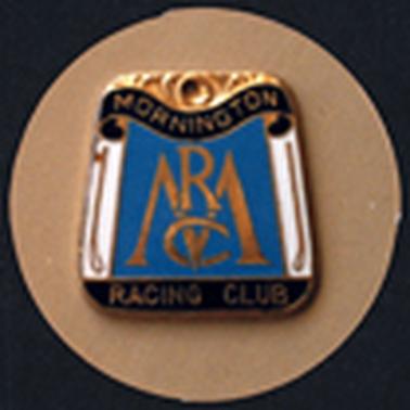 A rectangular metal badge, wider at the bottom, has a gold pattern at the top, ‘Mornington’ on a curved black ribbon below it and ‘Racing Club’ along the black strip at the bottom. In the centre is ’MRC’  in golden letters on a blue background, with a golden whip on either side of the monogram. The badge is photographed on a fawn  coloured circular background.