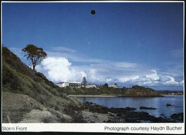 This is a view of the Royal Hotel from across the water, and it shows a threatening sky. It is taken from a grassy point across the water. There is a sloping hill to the left of the picture, with a solitary tree. Port Philip Bay and rocks in the foreground.