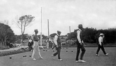 A bowling green is in the foreground of parkland. A group of four men is playing a game of bowls. Richard Edwards, in a hat, white trousers and a darker shirt, is playing with 3 others in dark trousers, white shirts with collar and tie and sleeveless vests. In the background are another group of bowlers, a couple on a park seat with a dog in front and a single bowler in suit and hat. 