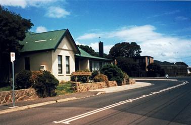 A double fronted, single storied house with pitched roof coloured green and cream coloured rendered walls. There is a mini verandah to the right of the front door. The house is set back from a low, stone fence at the front of the property. A sign in the front is indistinct in the photograph. A fairly newly surfaced road in front of the house curves to the left around it. 