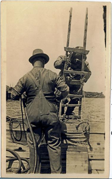There are two men at the edge of a wooden jetty. One, in a hat , shirt and overalls, stands with his back to the camera holding a long black pipe. The other in diving helmet and diver’s suit is on a ladder facing the camera preparatory to descending into the sea.