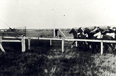 Photograph of a primitive race track, defined by a white post and single rail fence with a race in progress. A group of about 5 or 6 horses with jockeys on board is running from right to left. Two officials are standing in the background. On the left is a metal gate with four rails held open on top of a white, metre high post. Across a paddock there is a long line of trees in the distance. 