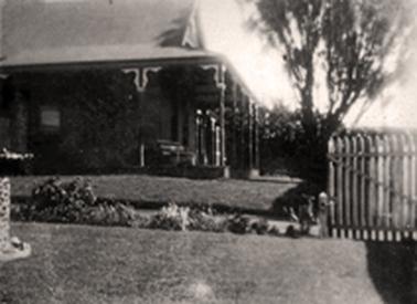 Single storied Edwardian house photographed from the side and showing the front verandah supported by decorated posts. The driveway is shown in the foreground between neat lawn edged with a garden bed. 