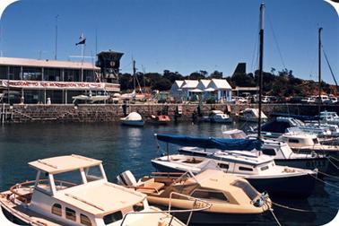 This shows a view of the Yacht Club from the water on the left of the photo. It is a two storey building with a lookout tower on the right at the end. There are verandas on both levels. On the top level there is a banner stretched across the entire length of the building. It says “Welcome to the Mornington Yacht Club”. The building has a brick coloured colourbond roof. In the centre of the photo is the little blue restaurant which has a distinctive roof It has four pitched sections. In the foreground there are several small boats. Some of the boats are sailing and some motor boats. Across the centre of the photo is the rock retaining wall. There is a man working on a yacht on the quayside. There are a couple of flag poles in view too. The sky is a deep blue. There is a glimpse of the obelisk on the hill on the right.
 




