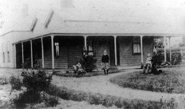 Single storied timber house in Federation style with metal double pitched roof and verandah. There is a sashed window on each side of the front door and more windows on the left. The verandah runs on the left side to a room at the back of the house. A lean-to at the back is just visible. Alfred Downward is seated on the right with daughter Ivy on his knee, Herbert is standing in the centre and Elsie is seated on the left. (Almost identical to item 1267.1)