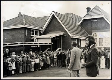 This image shows a large group of children standing in front of the school. The school is a brick building which has a pitched roof in 4 sections. It has large windows with white painted frames. The windows have small sections of glass.  Several female teachers can be seen among the pupils. In the foreground in front of the children, are two males. The man on the right has long dark hair, and a moustache.