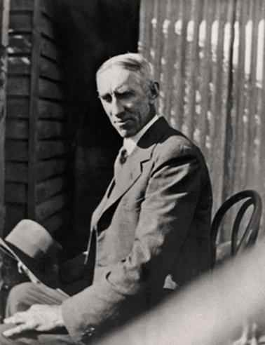 Man seated on a ‘bentwood’ chair in front of screen (? corrugated iron) beside a corner of a weatherboard building. He is dressed in a suit with shirt and tie and is holding a Trilby hat in his right hand and his left rests on a knee. His hair is thinning  and partly grey.