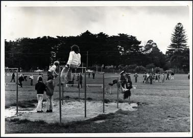 This image shows a playing field with lots of children playing. There is a climbing frame in the centre of the photo, and three children are climbing on the frame. In the background there are a line of Cyprus trees, and on the right of the photo is a Norfolk Pine treet.