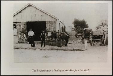 This image shows a corrugated iron building with a pitched roof, and large wooden doors. There are three gentleman in front of the building, alongside two horses. On the right of the picture are three carriages. 