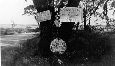 Photograph of twin tree trunk on road verge with unsurfaced road on the left. Three rather primitive signs are attached, one to each trunk and a third at the common base. The bodies of two foxes hang from a branch on the right from the trunk bearing a hand printed sign saying”Watch this tree for Jack Johnson’s 1944 catch”. The sign at the base says “Foxey’s Hangout Pride of TubbaRubba”. The sign on the left is illegible.