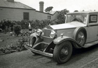 A Rolls Royce, c1930 and reg.no. AG666, stands in a driveway beside an extensive garden with stonework and a statue of a bird. A low0 hedge and the side of a weather board house are in the background.