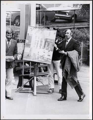 This photo shows 3 men. To the right is a man in a dark suit, walking with a coat over his arm. He appears to be talking and has a piece of paper in his left hand. To the right is another man, and he is holding a piece of paper with both hands. He has a patterned tie, dark double breasted jacket, and light trousers. The gentleman in the centre of the photo, is adjusting a large picture on a tripod easel. The picture appears to be of children playing.