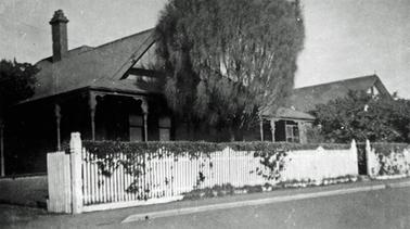 An Edwardian single storied house with verandah behind a white picket fence on the street.The house has a pitched roof with a single, tall chimney. There is a driveway on the left with open gates, a small pedestrian gateway to the right and a tall tree between house and fence. A shrub conceals what appears to be an extension of the house.